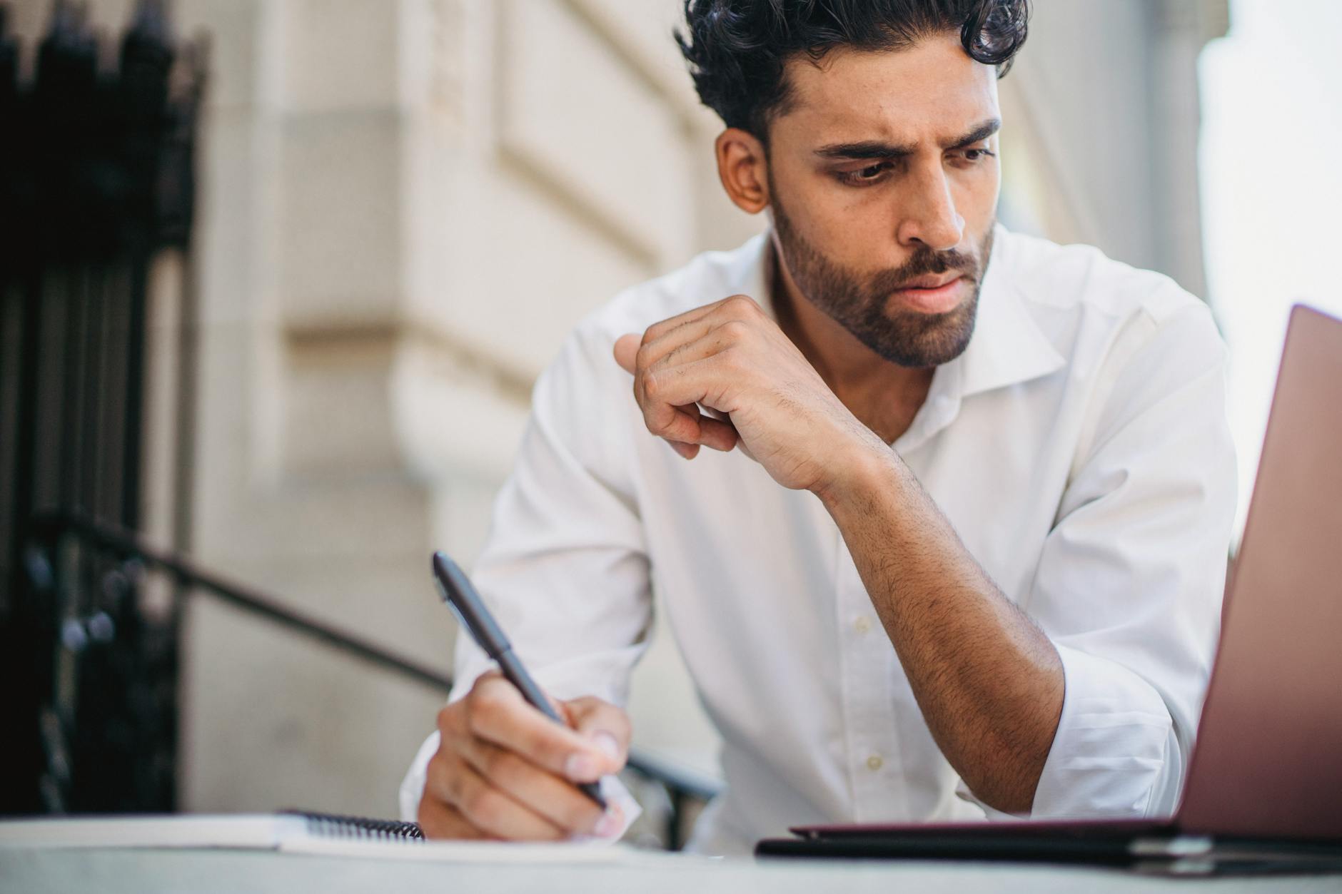 Skupiony mężczyzna w białej koszuli robi notatki przy laptopie na zewnątrz